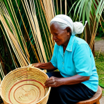 Home 9 An elderly Antiguan woman artisan, fully clothed in modest, comfortable traditional island attire, meticulously weaving a large straw market basket from dried palm fronds. Her hands are well-formed, showcasing years of skilled craftsmanship, with perfect anatomy and natural pose. She is seated on a low stool in an open-air workshop, bathed in soft, natural Caribbean sunlight, surrounded by bundles of raw materials and stacks of beautifully finished baskets. The background shows hints of lush tropical foliage. High-quality professional photography, detailed textures, natural colors, authentic scene, appropriate content, safe for work, family-friendly.
